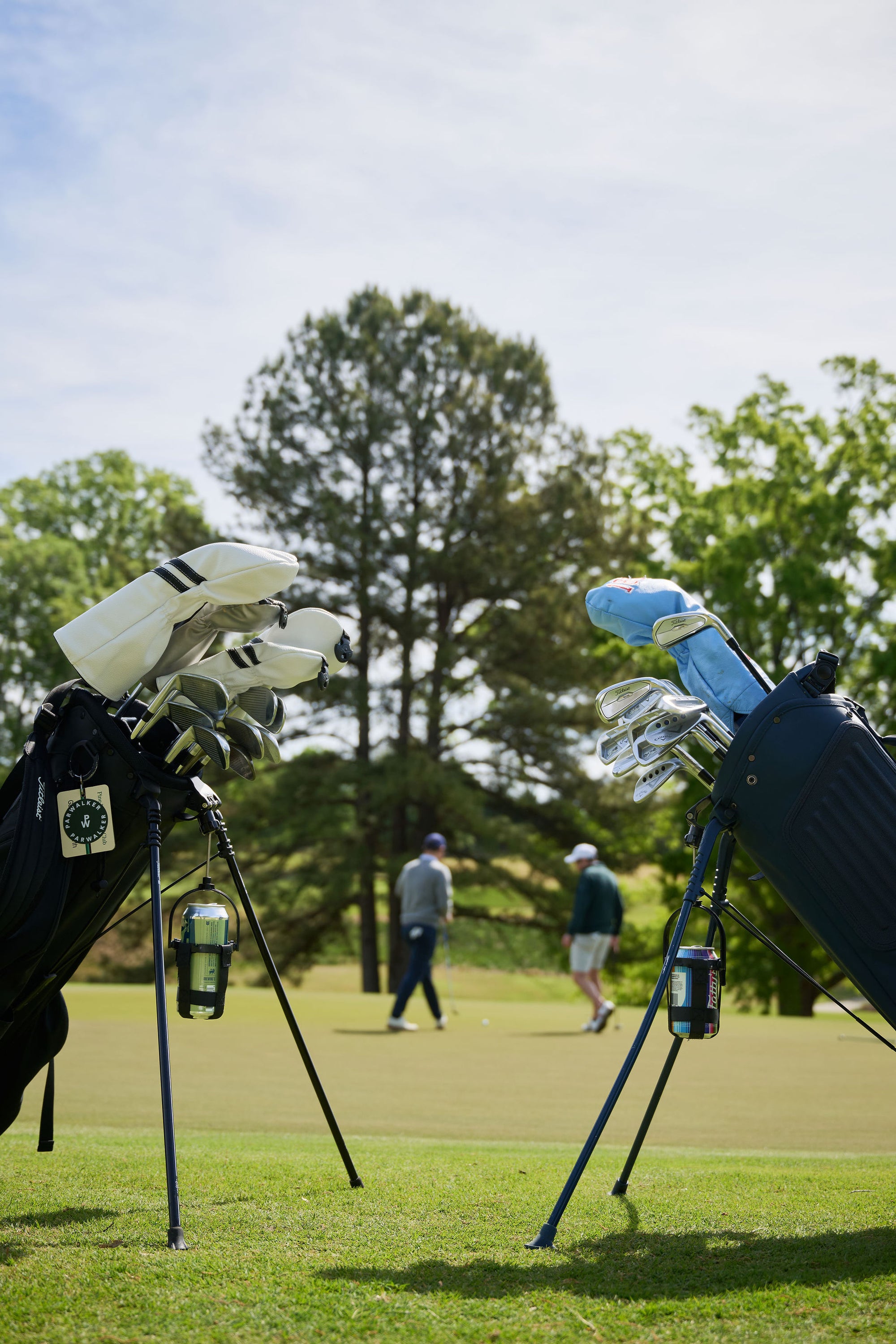 Two golf bags with Dangler drink holders attached, golfers walking a sunny course behind them.