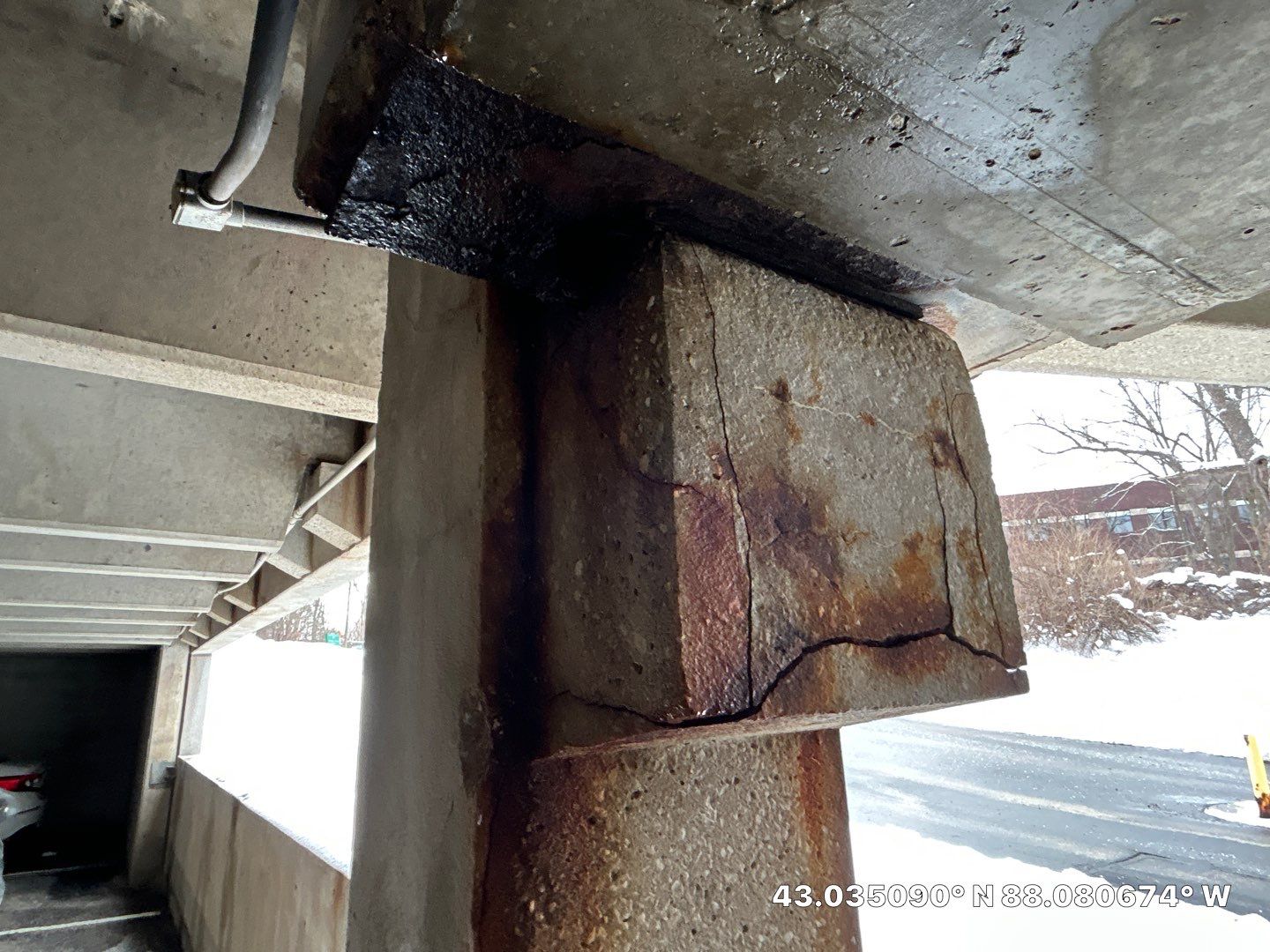 Severe freeze-thaw and corrosion-driven spalling at a parking garage beam-column connection with fractured concrete and heavy rust staining, photographed in winter conditions
