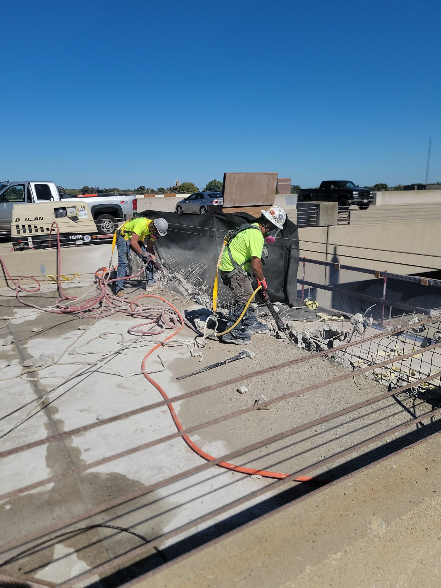 Two workers in safety vests spray-coat a concrete barrier wall on a sunlit parking ramp