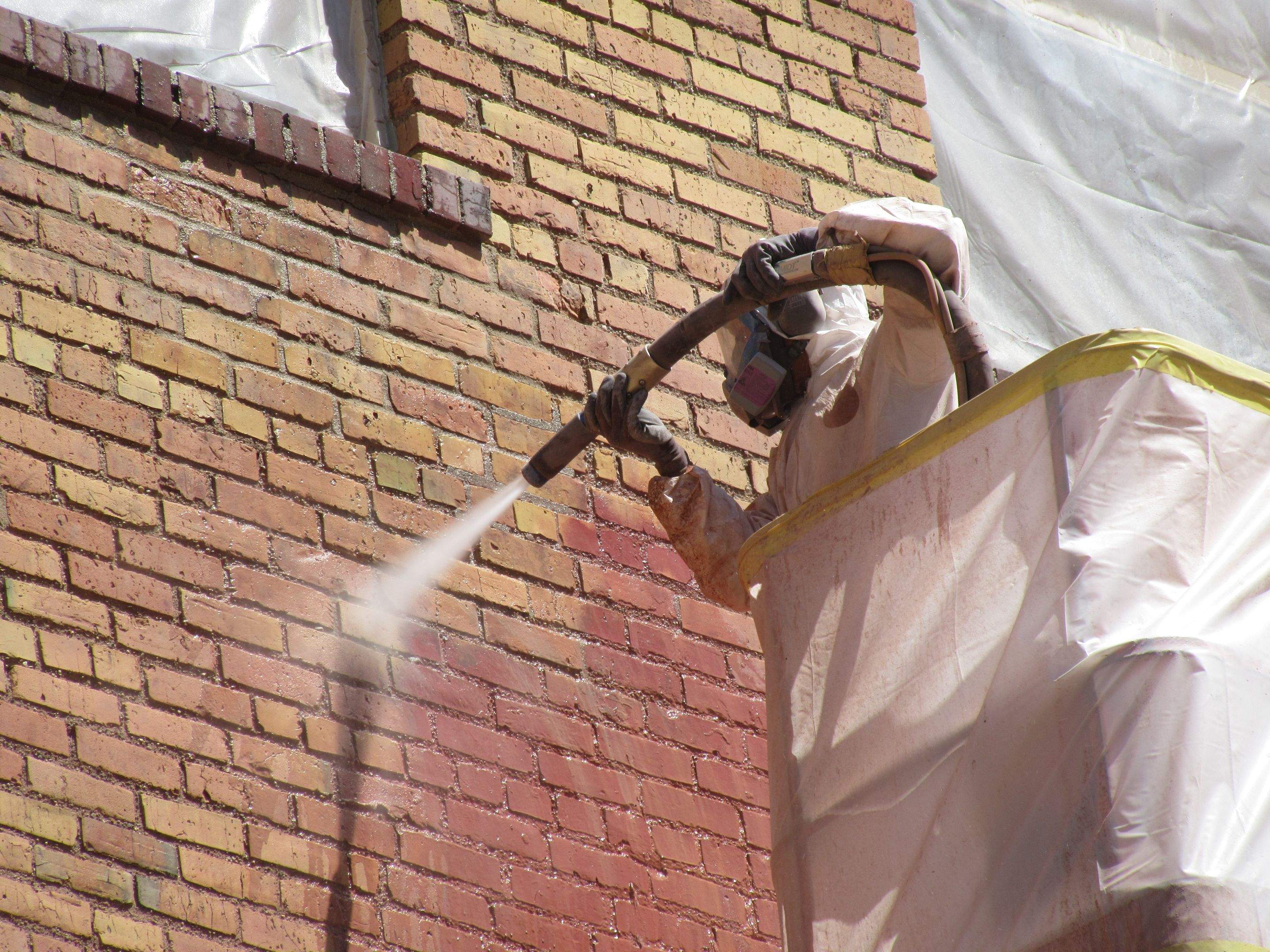 Close-up of an RSI technician in full PPE performing wet media blasting on the historic brick facade, with cleaned brick revealed below the still-painted upper section
