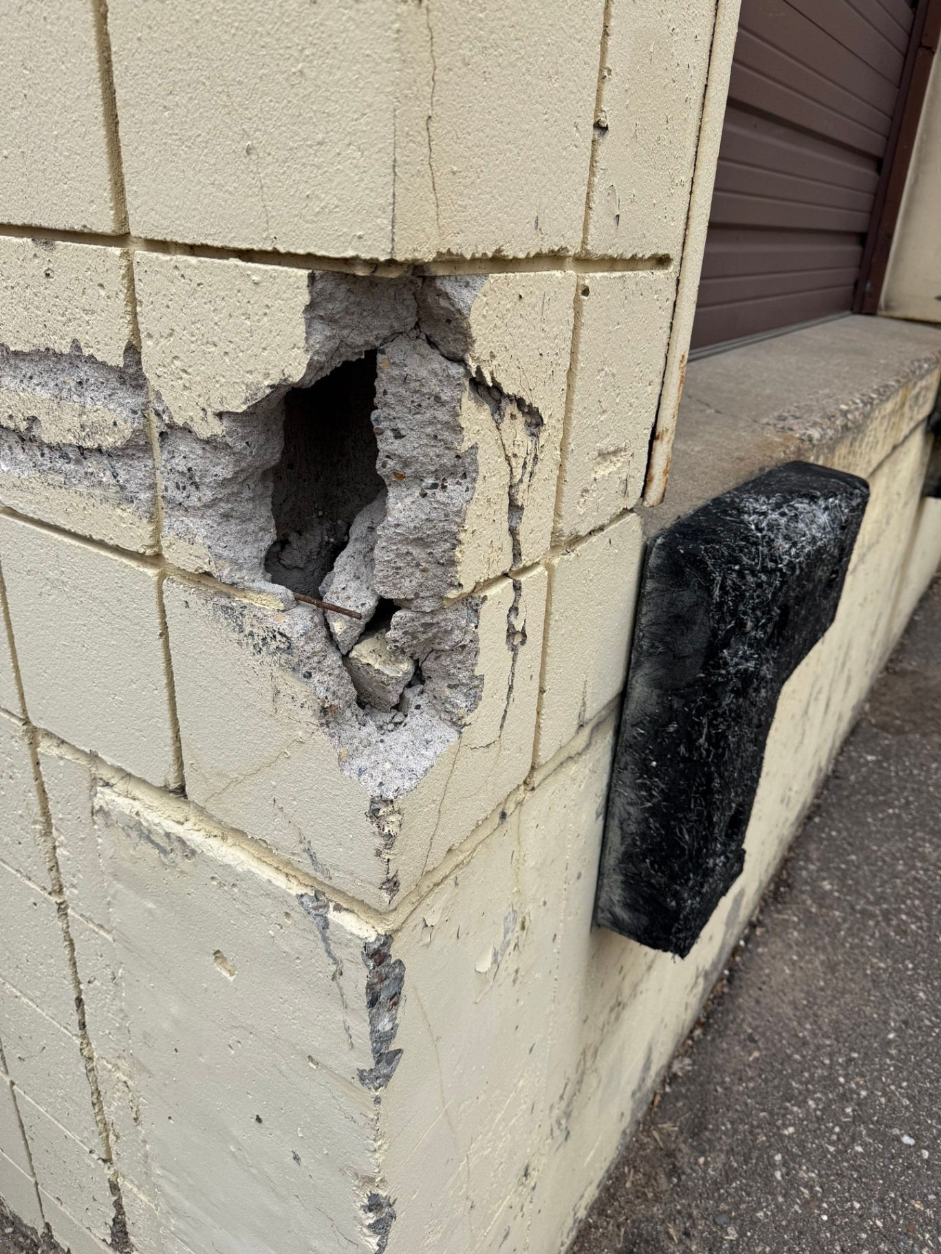 Severe spalling at the corner of a painted concrete masonry unit wall, with exposed aggregate and corroded steel reinforcement visible