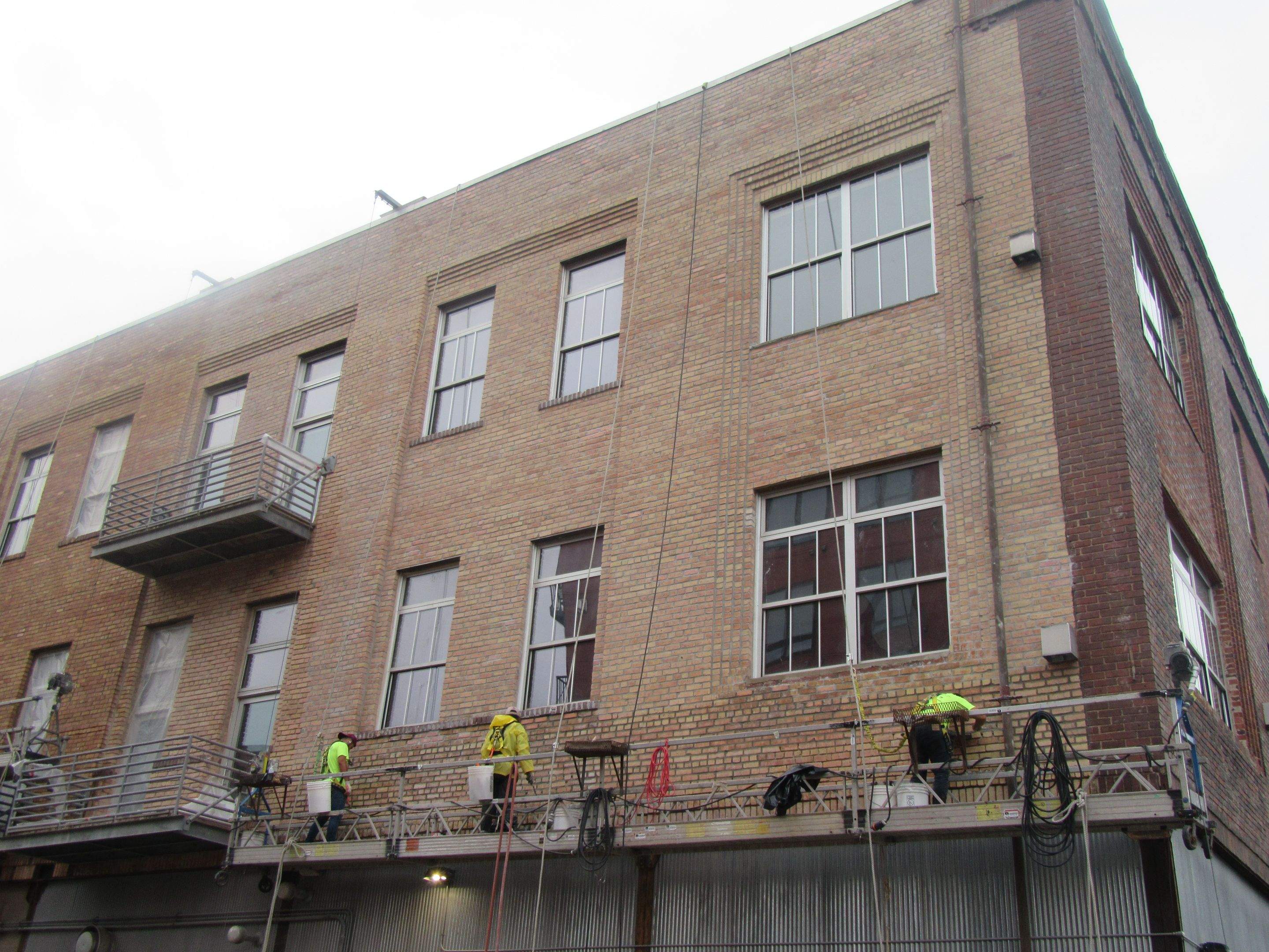 RSI crew in high-visibility safety gear working from a suspended swing-stage scaffold on the brick facade of the historic warehouse