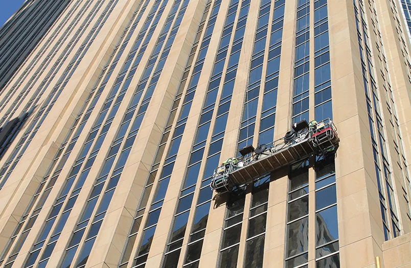 A two-point suspended scaffold (swing stage) hanging at mid-height down a downtown Minneapolis office tower facade, with reflective glass and stone visible behind the platform