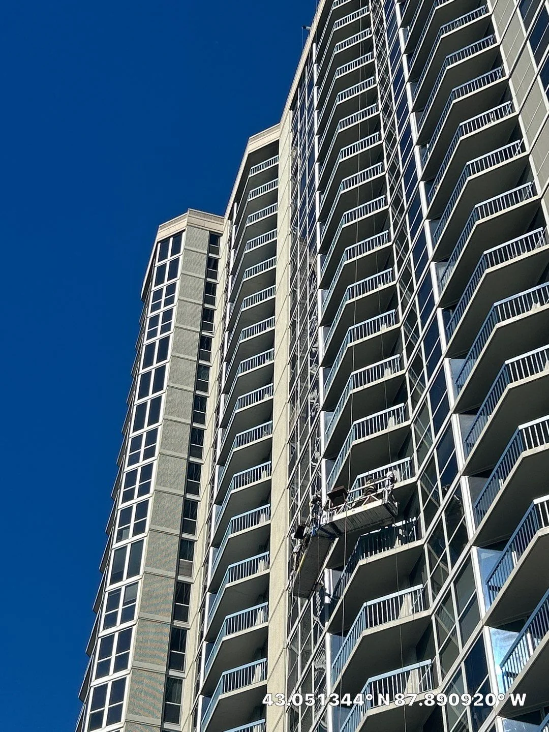Tall residential high-rise tower in Milwaukee with a two-point suspended scaffold (swing stage) hanging down the balcony facade against a deep blue sky