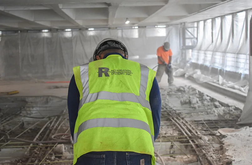 RSI crew member in safety vest inside an active parking-deck repair zone with reinforcing steel exposed across the slab floor