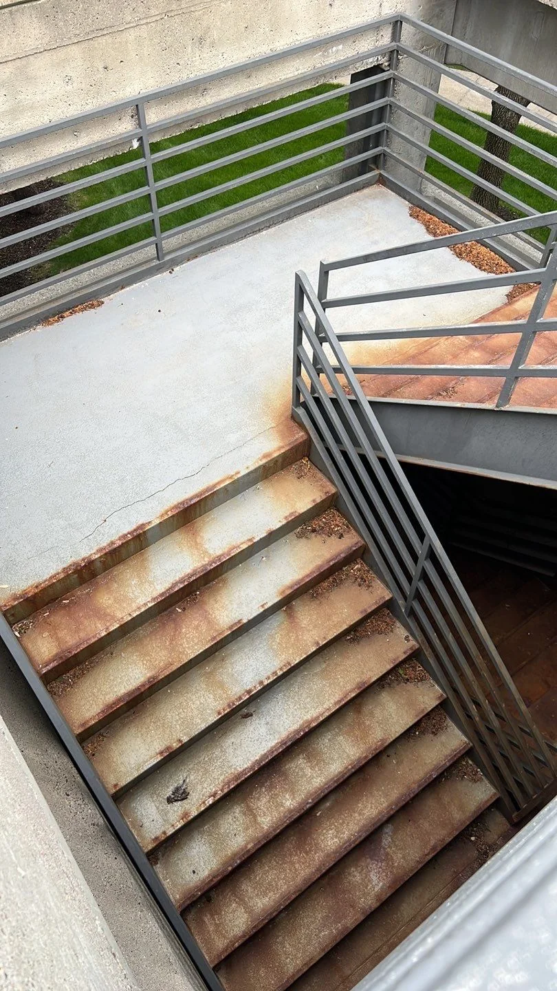 Corroded exterior steel stair with rust streaks running across the treads and stringers, leading down from a guard-railed landing